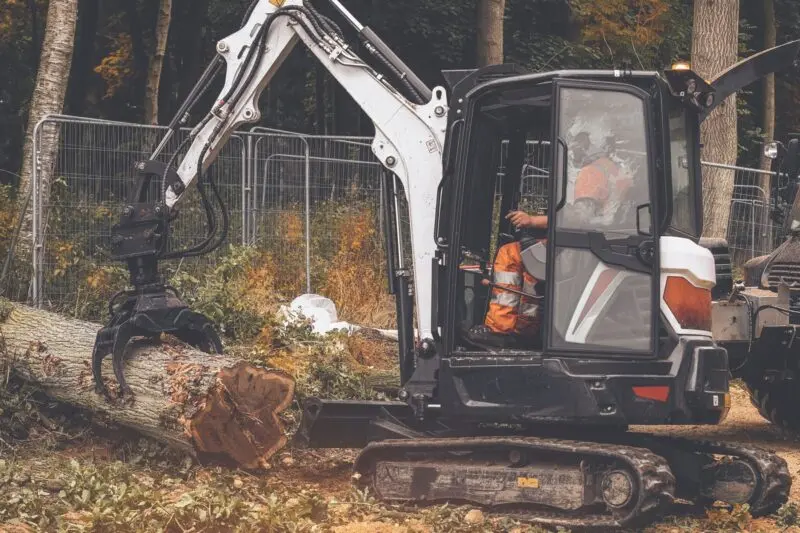 Excavator with a grapple lifting a felled tree in woodland